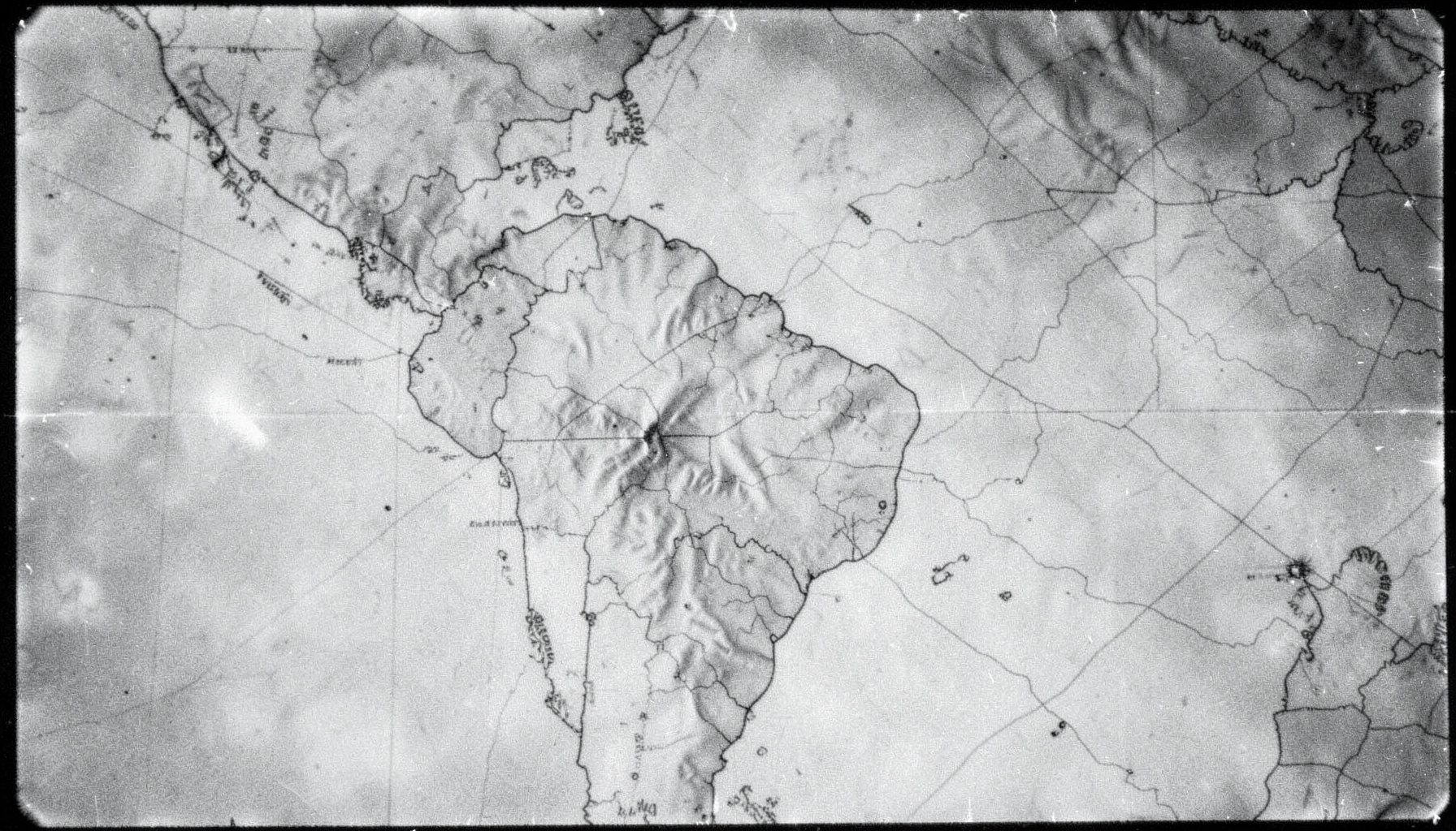 Fotografía analógica en blanco y negro de un mapa topográfico de Sudamérica con marcas sutiles, textura granulada y bordes ligeramente desgastados, iluminación lateral que resalta el relieve.